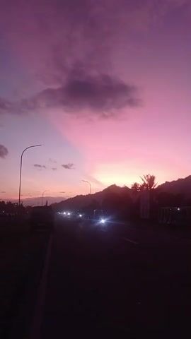 The video shows a scenic view of a road at dusk. The sky is a vibrant gradient of pink and purple, with some clouds scattered across it. The sun is setting behind a silhouetted mountain range in the distance. There are streetlights along the road, and several vehicles, including cars and motorcycles, are driving on it, their headlights illuminating the dark asphalt. Palm trees and other vegetation can be seen along the roadside, adding to the tropical atmosphere. The overall impression is one of a peaceful and beautiful evening.
