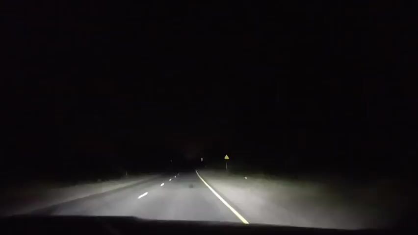 The video is shot from the perspective of someone driving a car at night. The headlights illuminate a dark, empty highway with a yellow line dividing the lanes. There are faint white markings on the road. On the right side of the road, a yellow triangular warning sign is visible. The surroundings are almost entirely black, indicating a lack of external light sources and possibly rural or undeveloped areas. The car appears to be moving forward steadily.