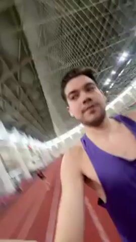 The video shows a man running on an indoor track. He is wearing a purple tank top and black shorts. He is holding the camera in front of him, so the video is a first-person perspective. The track is red with white lane markers. There is green turf in the center of the track. The building has a high ceiling and metal mesh along the top.