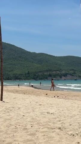 The video shows a sandy beach with people walking and swimming in the ocean. The ocean has waves, and there is a large, green, tree-covered hill in the background. The sky is blue and clear.