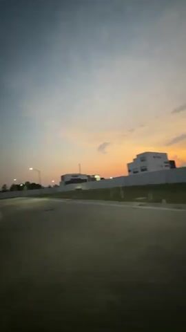 The video captures a view from a moving vehicle, likely a car, driving on a road during twilight. The sky transitions from blue to a warm orange glow near the horizon, indicating either sunrise or sunset. On the side of the road, there is a grassy field. In the distance, several buildings are visible, including one that appears to be under construction, with its brick structure exposed. Street lights line the road, and a white wall or fence runs along the perimeter of the area. A single white cloud floats in the sky.