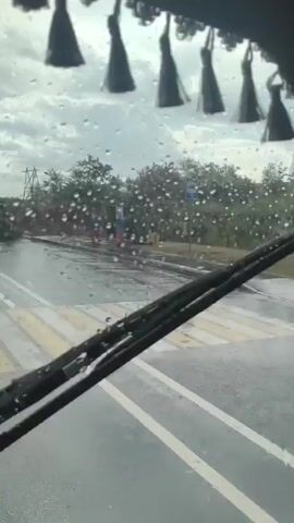 The video shows a view from inside a vehicle, looking forward. The windshield is wet, indicating rain, and there are decorative tassels hanging from the ceiling of the vehicle. The road ahead is wet and appears to be an asphalt surface with white lane markings. In the background, there are trees and vegetation. A structure with red, white, and blue columns, resembling a gate or checkpoint, is visible on the right side of the road. Further down the road, a large tanker truck has overturned and is lying on its side. The truck appears to be damaged, and its contents are unclear. The scene suggests a traffic accident has occurred, possibly due to adverse weather conditions. There are no people visible in the video.