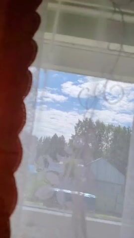 The video shows a view through a sheer curtain of a partly cloudy blue sky and some trees. In the distance, a white building and a blue tarp-covered object, possibly a pool, are visible. The curtain has a floral pattern.