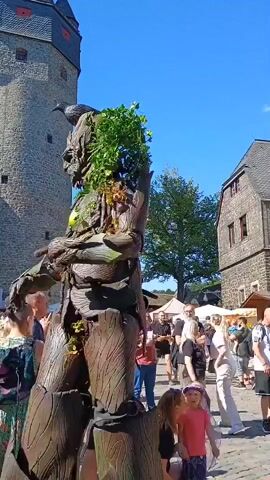 A costumed performer dressed as a large tree-like creature with greenery on its head walks through a town square filled with people. The performer is standing on stilts, making them appear very tall. The background shows historic stone buildings with timber framing and a tall stone tower. The sky is clear and blue, indicating a sunny day. People of various ages are watching the performer, some taking pictures.