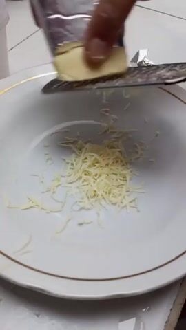 A hand with pale fingernails is grating a small block of butter into a white bowl rimmed with gold. The butter is wrapped in a piece of foil that protects the hand from the grater. The bowl is about a quarter full of shredded butter. The bowl sits on a white surface, likely a countertop.