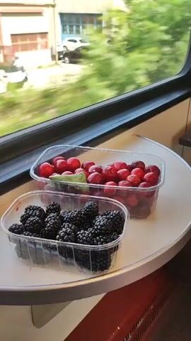The video pans across two clear plastic containers of berries sitting on a tray table by a train window. The container in the foreground holds blackberries, and the container in the background holds cherries. The window shows a blurred green landscape, indicating the train is in motion.