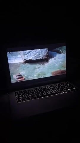 A laptop computer is shown in a dark room. The laptop screen displays a video of a waterfall. The laptop keyboard is illuminated.