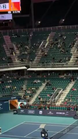 The video shows a tennis court stadium, with rows of green seats filled with people. The tennis court is blue, and there is a golf cart near the net. Banners with the names of sponsors are visible near the court. The stadium is well-lit.