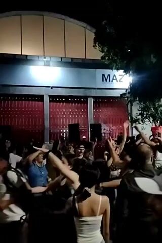 The video shows a lively street scene at night. A crowd of people are gathered outside a building with the word "MAZ" illuminated above a red-walled structure with visible roller doors. A man in a hat holds a cup aloft, and many others are raising their hands and clapping. Music is audible. The crowd extends down a street lined with trees and stalls. The street lights cast a warm glow. The scene suggests a celebration or event taking place outdoors.