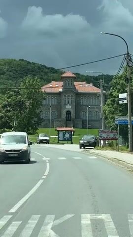 The camera is in a car, driving down a road in a town. The road is paved with a single white line in the middle. On the side of the road, there are light poles and power lines. Across the road, there is a large, stone building with a red tile roof and a small tower. There is a small bus stop in front of the building. There are several cars parked on the side of the road. There are trees and a grassy hill in the background. The sky is overcast.