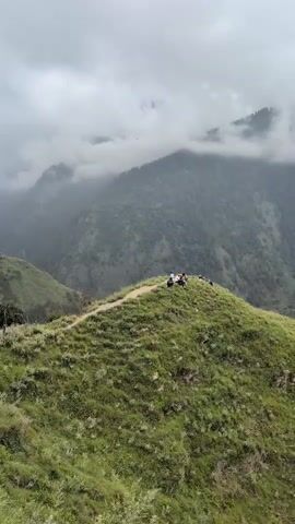 The video shows a mountainous landscape under a cloudy sky. A narrow dirt path winds along the crest of a grassy hill. On the path, several people are visible. On the right side of the path, closer to the camera, a group of people appear to be resting or gathered, with some sitting down. Further along the path, on the left side of the camera, a few more individuals are standing and observing the surroundings. The background features lush green slopes of other mountains, partially obscured by mist and clouds, creating a dramatic and atmospheric scene.
