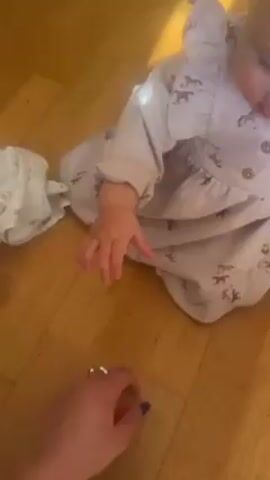 A woman's hand holds out a red crescent shaped toy. The toy has magnets on its flat end. A baby sitting on a wood floor reaches for the toy and grabs the toy from the woman's hand. The baby pulls the crescent shaped toy apart from a rainbow arc toy it had been attached to with magnets. The baby hands the red toy back to the woman. The woman says "Merci."