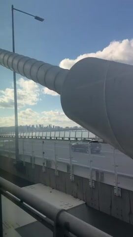 The camera captures a view from a bridge, moving slightly. A large white cylindrical structure dominates the left side of the frame, its scale implying it is part of the bridge's infrastructure. To the right of the structure is the bridge's road, with a barrier made of clear panels. In the distance, a cityscape is visible across a body of water, presumably a river or bay. The sky is blue with puffy white clouds. The clip captures a sense of travel and observation, with the focus on the bridge's construction and the distant urban panorama.