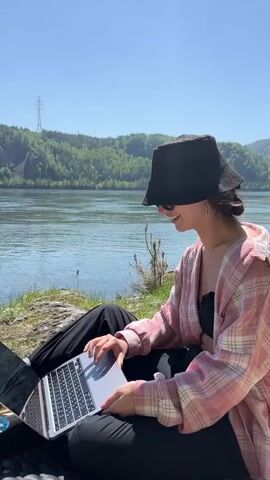 A young woman is shown sitting outdoors on a sunny day, using a silver laptop. She is wearing a black bucket hat that shades her face, sunglasses, a light pink and white plaid button-up shirt (open), a black top underneath, and black pants. Her right hand is on the laptop's trackpad. She is smiling and appears relaxed.

The setting is a scenic natural environment beside a wide body of water, likely a river, with green-blue water. Across the river, there are forested hills or mountains. A power line pylon is visible in the distance on the opposite bank. The sky is clear blue, indicating bright daylight. The woman speaks in Russian, saying "I'm hugging with the laptop here," suggesting she is comfortable and engaged with her work or activity. The location context provided is Sliznevo, Russia. There is no visible text on the laptop screen or elsewhere in the scene.