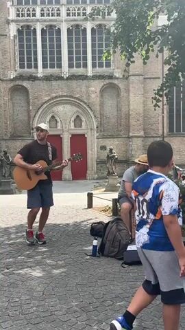 Two men are busking in front of a large, old brick building. The man on the left is wearing a black t-shirt, blue shorts, a white baseball cap, and sneakers. He is playing an acoustic guitar and appears to be singing. The man on the right is wearing a grey t-shirt, shorts, sandals, and a straw hat. He is seated on a Cajon, a type of percussion instrument, and appears to be drumming. A backpack, a water bottle, and a guitar case are on the ground between them.

The building behind them is made of brick with large, arched windows and ornate architectural details. A red door is visible in the center of the facade. The ground is paved with cobblestones. A tree with green leaves shades part of the scene. In the background, other people are visible, some sitting, and one walking towards the musicians. The lighting suggests it is daytime, likely sunny given the shadows. The overall atmosphere appears casual and relaxed, typical of street performers in a public square.

A sign on the Cajon reads "We're playing here! Your tips keep us going!" The text is in English. The scene appears to be set in Bruges, Belgium, given the architectural style.