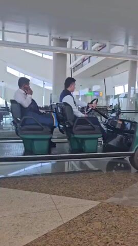 A green, six-seater golf cart is being driven down a wide, modern airport concourse. The cart has black seats and black luggage racks on the back. There are three people visible in the cart: a driver, a passenger in the front seat, and a passenger in the second row. The driver is wearing a white shirt. The passenger in the front seat is wearing a black and white shirt. The passenger in the second row is wearing a white shirt. The concourse has a polished marble floor, large windows, and a curved, illuminated ceiling. There are escalators on the left side of the cart. Several other people are visible in the background, walking or standing.
