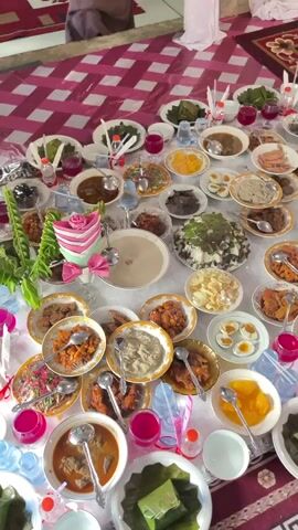 The video shows a large spread of food laid out on a table, likely for a gathering or celebration. The table is covered with a white tablecloth adorned with a pink and white checkered runner. Various dishes, served in bowls and on plates, are arranged closely together. These dishes include what appear to be curries, rice dishes, vegetable preparations, boiled eggs, and possibly desserts. There are also drinks in small glasses and bottles, some of which are red in color. Decorative elements such as pink flowers and green foliage are interspersed among the food. The scene suggests a communal dining experience, with a focus on a variety of food items. The background shows a patterned rug and draped fabric, indicating an indoor setting. The camera angle is overhead, providing a comprehensive view of the entire table arrangement.