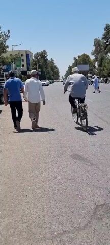 Two men walk side by side on a paved road, followed by a third man riding a bicycle. They are on the right side of the road, with traffic moving in the opposite direction. Several cars are visible on the road, including a white sedan and a dark-colored SUV. Trees line the sides of the road, and buildings can be seen in the background. The sky is clear and blue, indicating a sunny day. The scene appears to be in an urban or suburban environment.