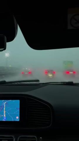The video shows the interior of a car driving in heavy rain. The windshield wipers are active and the visibility outside is very low due to the weather conditions. The car's navigation screen is visible in the foreground, displaying a map with a blue route highlighted. Several cars can be seen ahead through the rain-streaked windshield, their taillights glowing red. The overall atmosphere is gloomy and wet, typical of driving in a downpour.