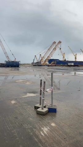 The video shows a rainy scene at a shipyard. In the foreground are three metal posts supported by concrete bases, wrapped with red and white caution tape. The ground is wet, with puddles reflecting the sky and objects around them. In the background is a large ship, dark blue and red, with the name "LEON KOSTECK" visible. Several yellow cranes are positioned around the ship, some of which are actively working, likely loading or unloading cargo. Further back are buildings, appearing to be offices or storage facilities associated with the shipyard. The sky is overcast with dark clouds, suggesting recent or ongoing rain. The overall atmosphere is industrial and somewhat bleak due to the weather.