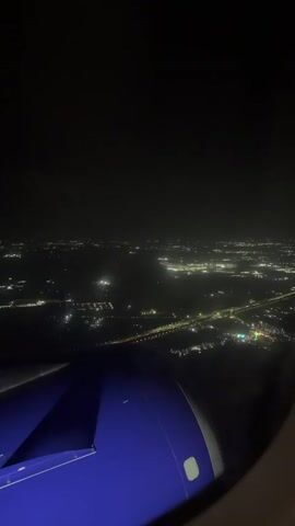 The video shows an aerial view of a city at night from an airplane window. The city lights twinkle below, forming patterns of urban sprawl. A highway with illuminated cars is visible, cutting through the landscape. Part of the airplane's wing and engine are in the foreground, illuminated by the ambient light. The sky is dark and appears to be cloudy.