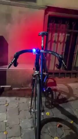 The video shows a blue bicycle with black handlebars and frame, standing on a cobblestone street, possibly at night. The bicycle has lights on the front that change colors from red to green to blue to orange. There is a red light illuminating the wall and gate behind the bike. The overall lighting in the scene is dim.
