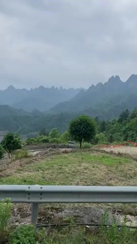The video shows a view of a mountainous landscape and a white house. The camera is positioned from a moving vehicle behind a guardrail. The landscape features rolling hills and distinctive rock formations in the distance, all under an overcast sky. In the foreground, there's a field of dry grass with a small tree. As the vehicle moves, the view shifts to show the side of a white house with a gray tiled roof. The house is set on a slightly elevated terrain with green vegetation growing around it. The camera continues moving past the house, showing more of its white walls and windows.