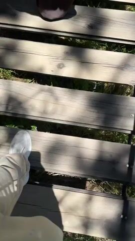 The video shows a first-person perspective of someone walking across a wooden bridge. The bridge is made of dark wooden planks with gaps between them, and sunlight filters through the trees, casting shadows. Green foliage is visible on either side of the bridge. The person is wearing light-colored pants and white sneakers. As the person walks, the camera pans slightly to show a small waterfall and a stream flowing beneath the bridge, surrounded by lush green vegetation and trees. The video ends with a close-up of the wooden planks of the bridge.