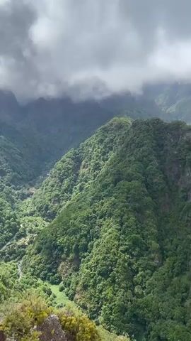 The video begins with a wide shot of a lush, green mountain range shrouded in clouds. The mountains are densely covered with trees, and the terrain appears rugged and steep. As the video progresses, the camera pans across the landscape, revealing more of the mountainous terrain and the thick vegetation. The lighting suggests an overcast day, with patches of sunlight breaking through the clouds in some areas. Towards the end of the video, the camera zooms out slightly, and two people's hands come into view in the foreground, appearing to rest on a railing or barrier. The backdrop remains the expansive mountain scenery, with a glimpse of a village nestled in a valley in the distance, and further back, a body of water can be seen.