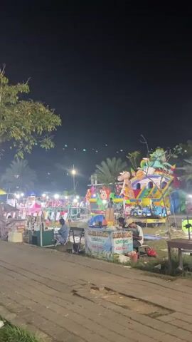 The video shows a night-time outdoor scene, possibly a fair or market. There are several stalls or attractions visible, including a colorful ride with what appears to be cartoon characters. Lights are present, illuminating the area. People are walking and gathered in groups. Some are seated near stalls. The ground is paved with interlocking stones, and grass is visible in some areas. There are trees in the background. The overall atmosphere appears to be festive.