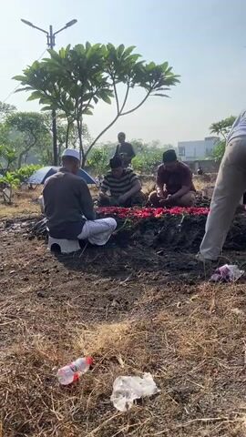 Several people gather around a freshly dug grave in an outdoor cemetery. Some are sitting around the grave, which is covered in red flower petals. Others are standing.  One of the standing men bends down and pours water on the grave. The environment includes sparse grass and several scattered discarded plastic items. A tree and a lamppost can be seen in the background. It is a sunny day.