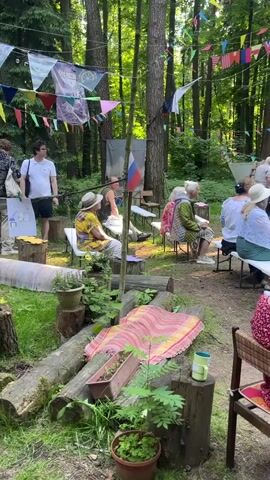 The video shows an outdoor gathering in a wooded area. Several people are seated on chairs facing a clearing where a large banner with historical images is displayed. A woman in a blue dress is standing in front of the banner and speaking into a microphone. Other individuals are visible in the background, some standing and some seated. Decorative flags are strung between trees above the gathering. The ground is covered with dirt and grass. In the foreground, there are logs and potted plants.