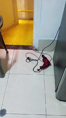 The video shows a red power drill with a black cord lying on a tiled floor.  A doorway leads to a yellow area with metal grating.  A person's hand reaches up to adjust a green metal grate above the doorway.  A light switch is visible on the wall next to the doorway.  A gray refrigerator stands along the wall on the right side of the frame.