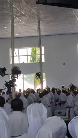 The video captures an indoor religious gathering or ceremony, likely a church service, taking place in a large, brightly lit hall. A substantial group of individuals, primarily adults, are visible, uniformly dressed in long white robes or garments. A distinctive feature is that many, particularly men, are adorned with ornate gold-colored crowns or headpieces.

The participants are observed in various states of reverence: some are standing, while others are bowing their heads, kneeling, or bending forward, suggesting prayer or worship. The overall atmosphere appears solemn and unified, with a discernible sound of singing or chanting in the background.

The setting is characterized by plain white walls, a grid-patterned ceiling, and large rectangular windows along one side that allow ample natural light into the space. Through the windows, green foliage and other buildings can be glimpsed, indicating an outdoor environment and suggesting it is daytime. Equipment such as oscillating fans and cameras on tripods are visible in the foreground, suggesting the event might be recorded or that ventilation is needed. The location context provided indicates this event is taking place in Anantigha, Nigeria. No discernible text is visible within the frame.