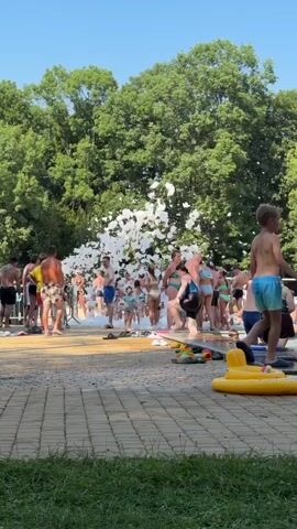 This media file shows a lively scene at an outdoor pool party, likely during a foam party. A large cloud of white foam billows from the center of the image, obscuring many of the individuals within it. Surrounding the foam are numerous people, dressed in swimwear, indicating they are enjoying the water and the festive atmosphere.

The subjects are primarily people of various ages, including children and adults, all engaged in the event. Scattered around the paved pool area are various objects, including inflatable pool toys like a yellow ring and a green alligator. The setting is a sunny outdoor location, characterized by a tiled or paved surface leading to a grassy area. Lush green trees form a dense backdrop against a clear blue sky, suggesting a warm, fair-weather day, likely in the late morning or afternoon.

The activity taking place is a foam party, a popular summer event where machines generate large amounts of foam for participants to play in. The people are actively interacting with the foam, with some fully immersed and others observing the spectacle. The overall mood appears to be one of fun and excitement, with a sense of collective enjoyment. The presence of a metal barrier on the left side might indicate a designated event area or queue.

There is no visible text in the media file. The location context of Krakow, Poland, suggests this event is taking place in a public or private recreational facility during the warmer months. The bright sunlight and clear skies are consistent with summer weather in Poland.