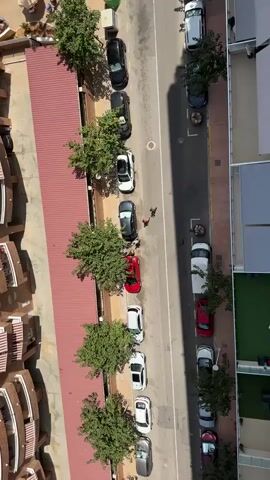 This is an aerial video clip from a drone looking down at a street with parked cars and buildings on either side. On the left, there is a long building with a red roof and several levels of balconies. Trees are planted along the edge of the building. On the right side of the street, there is a taller building and a sidewalk. The street itself is paved and lined with parked cars, most of which are white, black, and red. Two people can be seen walking on the sidewalk in the middle of the clip. The sun is shining, casting shadows. The scene appears to be in a warm climate, with palm trees visible in the background. The video is shot at an angle, giving a sense of depth and perspective.