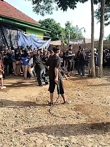 The video shows a group of people gathered in a dirt yard. The people appear to be participating in a cultural performance. Some of the individuals are wearing black clothing, and some are holding whips. There is a person on the ground near the center of the group. A person walks by, carrying an instrument or item, which looks like two drums strapped together. There is a building and wooden structure in the background. The area is under tree cover.