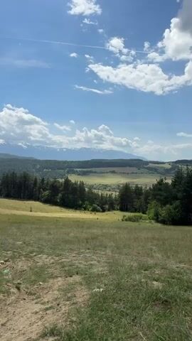 The video captures a scenic landscape under a bright, partly cloudy sky. In the foreground, there's a grassy field with a dirt path cutting through it. The midground features a dense line of evergreen trees. In the distance, a range of snow-capped mountains stretches across the horizon. The sky is predominantly blue, dotted with puffy white clouds, and there's a faint contrail visible. The overall impression is one of a peaceful and natural setting.