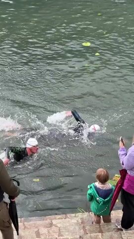 A man swims in a body of water, watched by three onlookers, including a child. The swimmer wears a black wetsuit and a white swim cap. They are in murky green water, creating splashes as they move. At least one lily pad is visible in the water. On the bank, a man in a vest and trousers holds a black umbrella. To his right, a woman in a purple jacket holds a red umbrella and films with her phone. A small child in a green crocodile-themed jacket stands between them. The group is positioned on stone steps leading down to the water. The overall environment seems to be outdoors, possibly a lake or river.