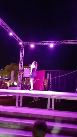 A woman is singing on a stage at night. She is wearing a black dress and white shoes. There are lights on the stage and in the background. There are also speakers on the stage. In the background, there is a large building with many lights. There are also people in the foreground watching the performance.