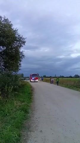 A fire truck with flashing blue and red lights drives down a dirt road towards the camera. Several people walk on the right side of the road in the same direction as the truck. A motorcycle passes the fire truck from behind on the right side of the road. The sky is cloudy and the surrounding environment appears to be a field with a tree on the left side of the road.