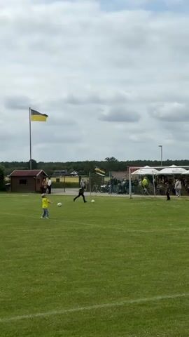 A full shot captures a green soccer field under a cloudy sky. In the foreground, a young boy in a yellow shirt and blue jeans approaches a soccer ball on the left. In the midground, another young boy in a black long-sleeved shirt and black pants kicks a soccer ball towards the goal on the right. Another soccer ball rests on the grass near the goal. In the background, spectators gather near a building with a red roof, a large black and yellow flag waving from a tall pole, and a cluster of white tents on the right. A dark green forested area is visible further back.