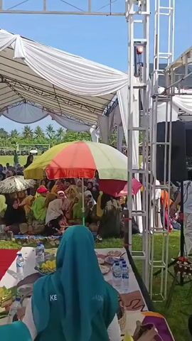 The video shows an outdoor event, likely a festival or gathering, taking place on a grassy field under a bright blue sky. Several white tents provide shade for attendees, and the perimeter is populated with various vendors and more people.  

In the foreground, individuals are seated on the grass, many wearing headscarves. A band is set up, consisting of musicians playing guitar and keyboard.  The camera focuses on them at various points, along with the audience. 

Various objects are visible, including a speaker, water bottles on a table, and a large umbrella. The atmosphere appears lively, suggesting a community event. The camera moves around, showing different perspectives of the scene and highlighting the various activities taking place.