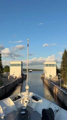 The video shows the bow of a white ship as it approaches a canal lock. Two white buildings flank the entrance to the lock. A blue flag flies from a pole on the ship. The sky is blue with scattered white clouds. Trees are visible on the right side of the frame. Another ship is visible in the distance on the water. The water is calm and reflects the sky.