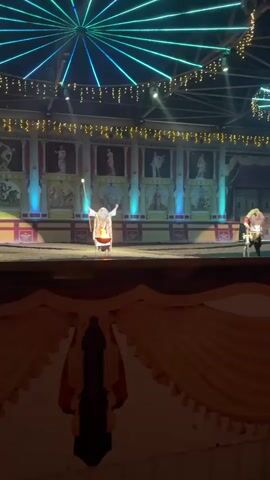 A person in a costume that resembles an ancient wizard or priest stands on a dirt stage in front of a muraled backdrop. The person has a long white beard and wig, and wears a golden robe adorned with patterns. They are holding a staff with a glowing tip in their left hand and are gesturing with their right hand over a fire pit in front of them. The fire pit is burning brightly. The backdrop features painted scenes of classical figures and architecture. Above the backdrop, decorative string lights and a circular light fixture with green and blue lights are visible. In the foreground, there is a draped fabric, possibly part of a stage curtain. The overall scene suggests a theatrical performance or reenactment.