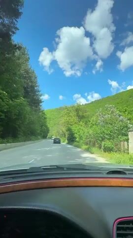 A driver is on a road in a mountainous, forested area. The trees on either side of the road are green.  The sky is bright blue with scattered white clouds. A dark car is ahead of the driver.