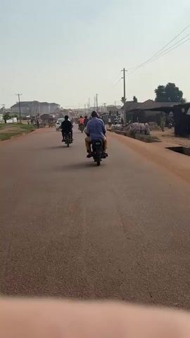 A video taken from a moving motorcycle shows a dusty, paved road with several other motorcycles and a three-wheeled vehicle. The person in the foreground is wearing a blue shirt and yellow pants, riding on the back of a motorcycle. In front of them, another motorcycle with two riders is visible. Further down the road, there are more motorcycles, cars, and people walking. Buildings and stalls line the sides of the road, and there are piles of goods, possibly sacks of produce, on the right side. The sky is clear and the sun is shining.