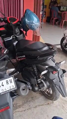 The video shows a row of motorcycles parked outside a shop. In the foreground, there is a black scooter with a blue helmet resting on its seat. To the right of the black scooter, there is a white scooter. Further back, more motorcycles are visible, including a red one and a blue and black one. The shop's interior can be seen through an open doorway, where a person is sitting at a computer. There are posters and banners displayed on the shop's wall. The ground is concrete and somewhat dusty.