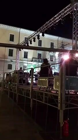 A video shows a night-time outdoor event with a stage setup. The stage is constructed from metal scaffolding and features lighting equipment and speakers. Several people are visible on and around the stage, appearing to be setting up or operating equipment. In the background, a building with multiple windows is illuminated, suggesting the event is taking place in a town or city square. A red fire extinguisher is mounted on a metal pole in the foreground. The lighting is dramatic, with spotlights illuminating parts of the stage and creating shadows. The overall impression is of a prepared event, possibly a concert or performance, in its early stages of setup.