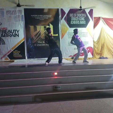Two dancers perform on a stage in front of event banners. The dancer in the purple jersey with the number 34 is on the right. The dancer in the black outfit is on the left. The stage is lit with purple light. A metal staircase is on the right side of the video. A ceiling fan is in the upper left corner of the scene.